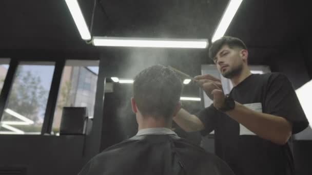 魅力的な男性は、理髪店で近代的なヘアカットを取得しています。床屋はスプレーで髪を濡らし、くしをします。ハンドヘルドショット。4k. — ストック動画