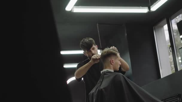 床屋ははさみでクライアントの髪を切ります。魅力的な男性は、理髪店で近代的なヘアカットを取得しています。ハンドヘルドショット。4k. — ストック動画