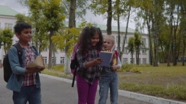 Okula geri dön. Sırt çantaları ile üç arkadaş sarılma ve okul önünde gülüyor. Okul bahçesinde eğlenen Okul Çocukları Karışık Irk Grubu. Yavaş Çekim Çekimi.