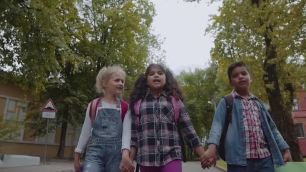 Drie vrienden met rugzakken gaan naar school. Gemengde raciale groep van school kinderen lopen langs de straat handen vasthouden. Slow Motion-opname. — Stockvideo