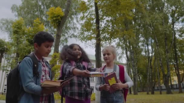 Tillbaka till skolan. Tre kompisar med ryggsäckar som huging och skrattar framför skolan. Blandad ras grupp skolbarn ha kul att kasta upp böcker i skolgården. Slow motion shot. — Stockvideo