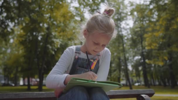 Bliska mała piękna uczennica rysunek z kolorowych ołówków, siedząc na ławce w słonecznym parku. Strzał w zwolnionym tempie. — Wideo stockowe