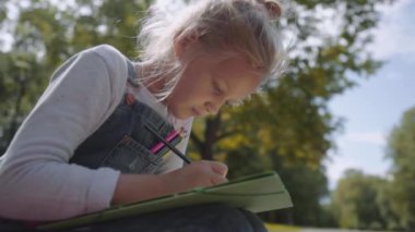 Sunny Park bir bankta oturan renkli kalemler ile çizim küçük güzel okul kız yakın. Yavaş hareket çekimi.