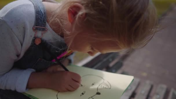 Cerrar Pequeña niña hermosa escuela dibujo con lápices de colores, sentado en un banco en el parque soleado. Disparo de cámara lenta . — Vídeo de stock