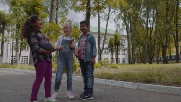 학교로 돌아갑니다. 배낭을 가진 세 친구는 학교 앞에서 포옹하고 웃고 있습니다. 학교 마당에서 재미를 학교 아이들의 혼합 인종 그룹. 슬로우 모션 샷. — 비디오