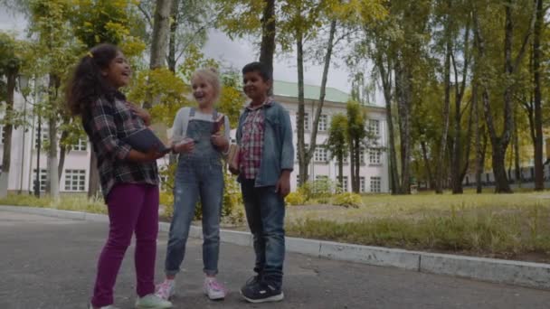 Je retourne à l'école. Trois amis avec des sacs à dos étreignant et riant devant l'école. Groupe racial mixte d'écoliers qui s'amusent dans la cour d'école. Prise de vue au ralenti . — Video