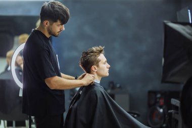 Berber yeni saç modeli berber dükkanı için müşteri hazırlanıyor. Sandalyede oturan ve aynaya bakan siyah bir elbise giyen adam. Hizmet ve saç kesimi kavramı.