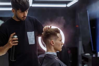 Berber kuaförde saç kesiminden sonra saç spreyi ile saç stili yapıyor. Genç yakışıklı beyaz adam modern bir kuaförde saçını kestiriyor..