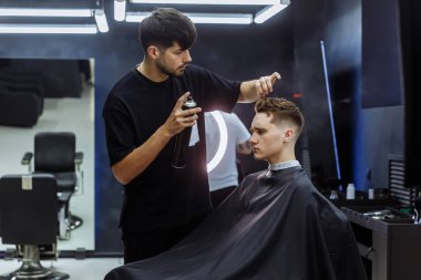 Berber kuaförde saç kesiminden sonra saç spreyi ile saç stili yapıyor. Genç yakışıklı beyaz adam modern bir kuaförde saçını kestiriyor..