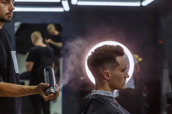 Барбер робить зачіску з розпилювачем волосся після стрижки в перукарні. Молодий гарний кавказький чоловік стрижеться у сучасному перукарні.. — стокове фото