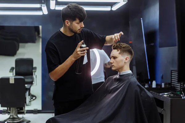 Berber kuaförde saç kesiminden sonra saç spreyi ile saç stili yapıyor. Genç yakışıklı beyaz adam modern bir kuaförde saçını kestiriyor..