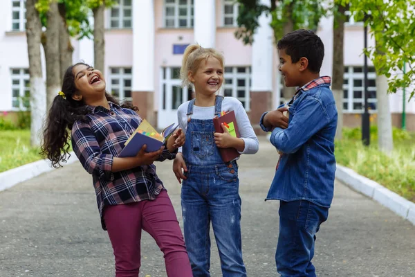 학교로 돌아갑니다. 배낭을 들고 학교 앞에서 포옹하고 웃으며 세 친구를 가까이에 두세요. 학교 마당에서 재미를 갖는 학교 아이들의 혼합 인종 그룹. — 스톡 사진