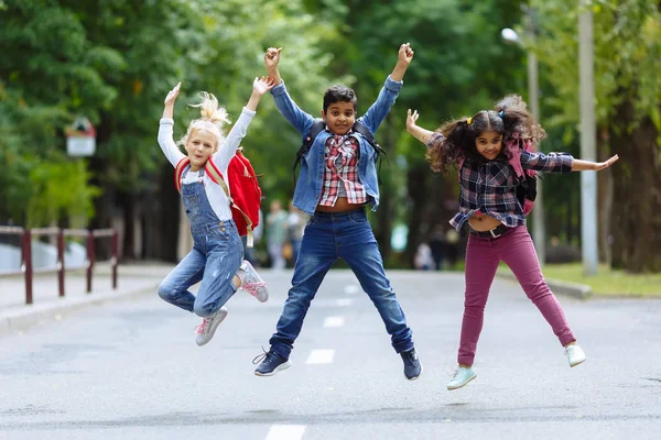Vegyes faji csoport boldog gyermekek Ugrás a parkban közelében iskola. Az alapfokú oktatás koncepciója. — Stock Fotó
