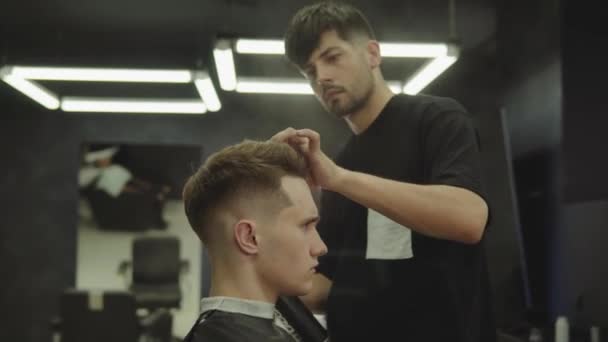 Peluquero hace el peinado con spray para el cabello después de corte de pelo en la peluquería. Joven hombre caucásico guapo consiguiendo un corte de pelo en una peluquería moderna. Un disparo de mano. 4K . — Vídeos de Stock