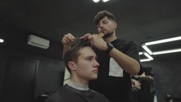Barbeiro corta o cabelo do cliente com uma tesoura. Um homem atraente está a cortar o cabelo na barbearia. Um tiro à mão. 4K . — Vídeo de Stock