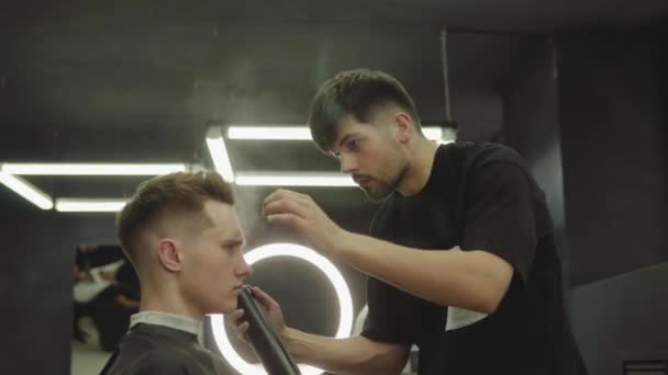 Barber fait coiffure avec de la laque après la coupe de cheveux au salon de coiffure. Jeune homme blanc beau se faire couper les cheveux dans un salon de coiffure moderne. Tir à la main. 4K . — Video