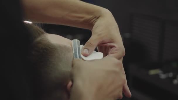 Açık boğazlı, keskin tıraş bıçaklı profesyonel berber erkek şakaklarında saç tıraşı olur. Çekici erkek berber dükkanında modern bir saç kesimi yaptırıyor. Yakın çekim. El kamerasıyla. 4K. — Stok video