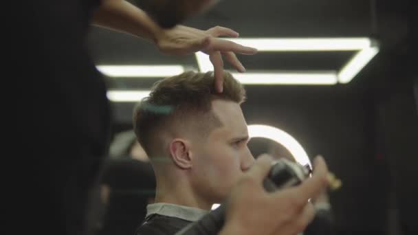 Peluquero hace el peinado con spray para el cabello después de corte de pelo en la peluquería. Joven hombre caucásico guapo consiguiendo un corte de pelo en una peluquería moderna. Un disparo de mano. 4K . — Vídeos de Stock