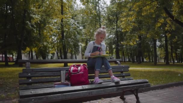 Malá krásná školní dívka s barevnými tužkami, která sedí na lavici ve slunném parku. Výstřel z pomalého pohybu. — Stock video