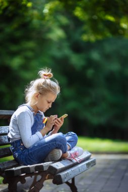 Happy Little güzel okul kız ilkokul çağında bir park bankında oturan ve yaz günü açık havada cep telefonu bakarak.