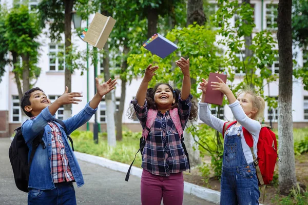 学校に戻れ学校の前で抱き合い、笑うバックパックを持つ3人の友人。校庭で本を投げる楽しみを持つ学校の子供たちの混合人種グループ. — ストック写真