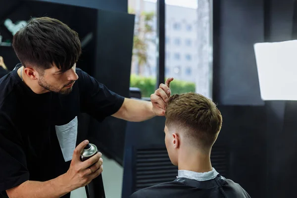 Barber makes hair styling with hair spray after haircut at the barber shop. Young handsome Caucasian man getting a haircut in a modern hairsalon.