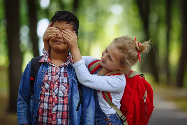 Two school Friends having fun in park. Caucasian Girl covering eyes to surprised afro american boy.