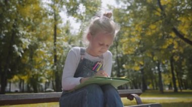 Sunny Park bir bankta oturan renkli kalemler ile çizim küçük güzel okul kız yakın. Yavaş hareket çekimi.