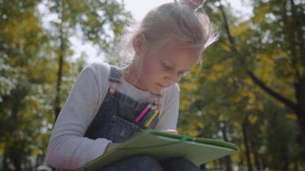 Close up Little beautiful school girl drawing with colored pencils, sitting on a bench in sunny park. Slow Motion Shot. — Stock Video