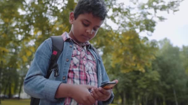 屋外の肖像画アフロアメリカの幸せな学校の男の子は、スマートフォンを持っています。携帯電話を見ている若い学生。学校の概念に戻る。スローモーションショット. — ストック動画