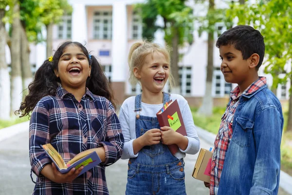 Torniamo a scuola. Chiudi Tre amici con gli zaini che si abbracciano e ridono davanti alla scuola. Gruppo misto razziale di bambini della scuola che si divertono nel cortile della scuola . — Foto Stock