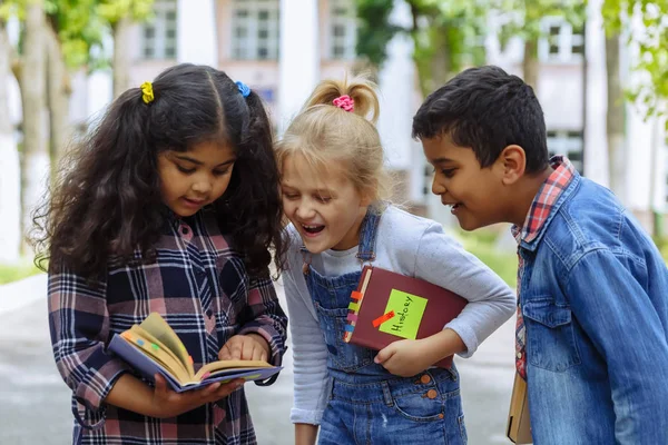 Tillbaka till skolan. Närbild tre vänner med ryggsäckar huging och skrattar framför skolan. Blandad ras grupp skolbarn ha kul på skolgården. Stockbild