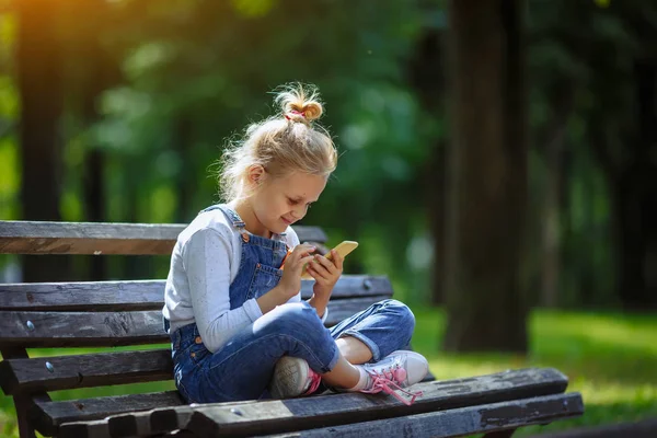 Happy Little güzel okul kız ilkokul çağında bir park bankında oturan ve yaz günü açık havada cep telefonu bakarak.