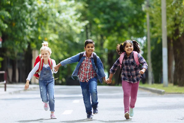 Okula dönüyoruz. Karışık ırksal grup sırt çantası ile mutlu İlköğretim öğrencileri el dışarıda tutarak çalışan. İlköğretim konsepti.