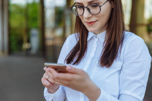 Portrait young business woman wearing white shirt using smartphone out doors. Female reading sms message in working process.