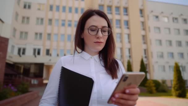 Μπροστινή θέα της επιχείρησης γυναίκα φορώντας λευκό πουκάμισο περπάτημα και χρησιμοποιώντας ένα έξυπνο τηλέφωνο σε ένα δρόμο της πόλης — Αρχείο Βίντεο