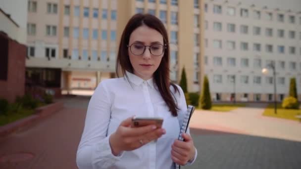 Vista frontal de la mujer de negocios con camisa blanca caminando y usando un teléfono inteligente en una calle de la ciudad. Disparo de cámara lenta . — Vídeos de Stock