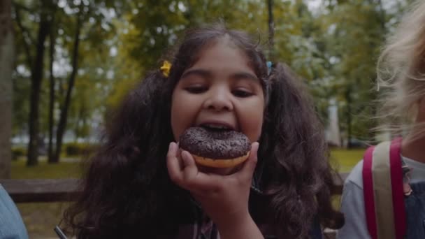 Petite écolière afro-américaine drôle mangeant un beignet au chocolat avec une émotion heureuse au parc . — Video