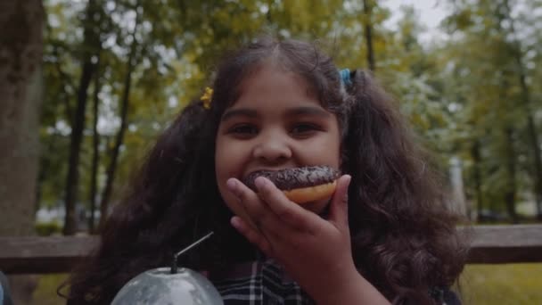 Poco divertido afro-americano schhoolgirl comer donut de chocolate con emoción feliz en el parque . — Vídeos de Stock