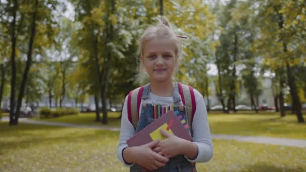 Colegiala alegre con mochila sosteniendo un libro que se queda en el patio de la escuela. Retrato de una chica muy inteligente sonriendo al aire libre. Disparo de cámara lenta . — Vídeos de Stock