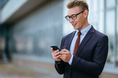 Mutlu işadamı, ofisin önünde duran bir mesaj okurken gülümsüyor. Açık havada akıllı telefon kullanarak trendy giysiler içinde Erkek icra yöneticisi. Modern teknoloji kavramı