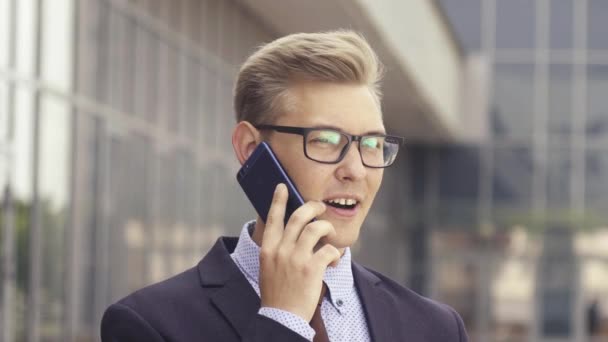 オフィスビルの前で携帯電話で話すメガネをかけた若いハンサムな白人ビジネスマン。屋外でスマートフォンを使用してトレンディな服を着た男性エグゼクティブマネージャー。現代技術の概念 — ストック動画