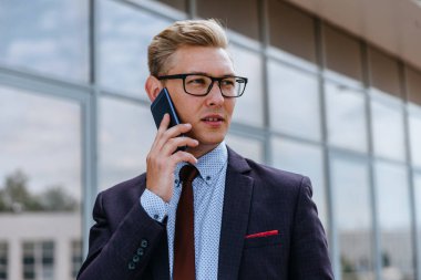 Gözlüklü genç beyaz işadamı ofis binasının önünde cep telefonuyla konuşuyor. Açık havada akıllı telefon kullanarak trendy giysiler içinde Erkek icra yöneticisi. Modern teknoloji kavramı