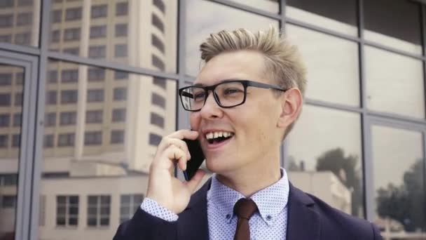 Jovem e bonito empresário caucasiano de óculos falando no celular em frente ao prédio de escritórios. gerente executivo masculino em roupas da moda usando smartphone ao ar livre. Conceito de tecnologia moderna — Vídeo de Stock