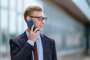 Gözlüklü genç beyaz işadamı ofis binasının önünde cep telefonuyla konuşuyor. Açık havada akıllı telefon kullanarak trendy giysiler içinde Erkek icra yöneticisi. Modern teknoloji kavramı