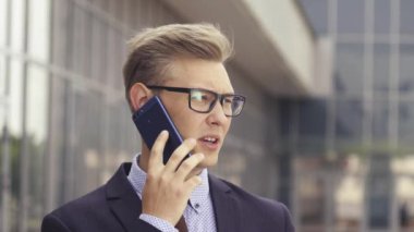 Gözlüklü genç beyaz işadamı ofis binasının önünde cep telefonuyla konuşuyor. Açık havada akıllı telefon kullanarak trendy giysiler içinde Erkek icra yöneticisi. Modern teknoloji kavramı
