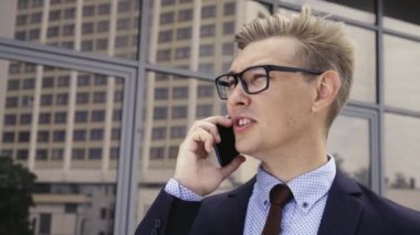Gözlüklü genç beyaz işadamı ofis binasının önünde cep telefonuyla konuşuyor. Açık havada akıllı telefon kullanarak trendy giysiler içinde Erkek icra yöneticisi. Modern teknoloji kavramı
