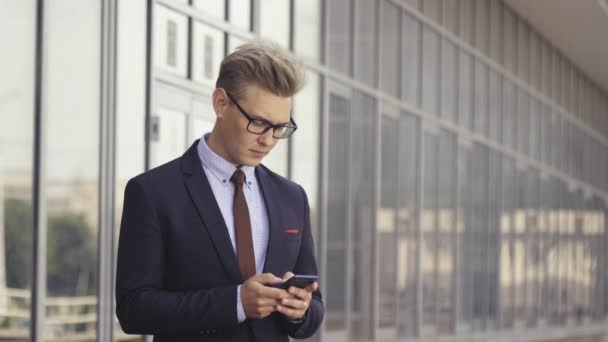Happy businessman smiling as he reads a text message standing outside the office. Male executive manager in trendy clothes using smartphone outdoors. Modern technology concept — Stock Video