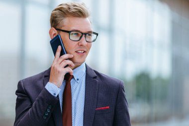 Gözlüklü genç beyaz işadamı ofis binasının önünde cep telefonuyla konuşuyor. Açık havada akıllı telefon kullanarak trendy giysiler içinde Erkek icra yöneticisi. Modern teknoloji kavramı