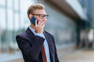 Gözlüklü genç beyaz işadamı ofis binasının önünde cep telefonuyla konuşuyor. Açık havada akıllı telefon kullanarak trendy giysiler içinde Erkek icra yöneticisi. Modern teknoloji kavramı